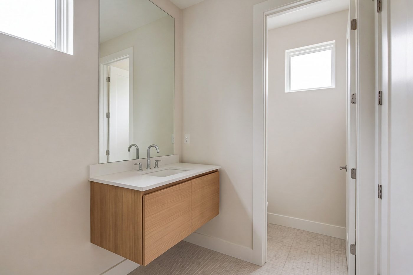Small hall bathroom with pale walls, a wood vanity and a tall mirror reflecting window light